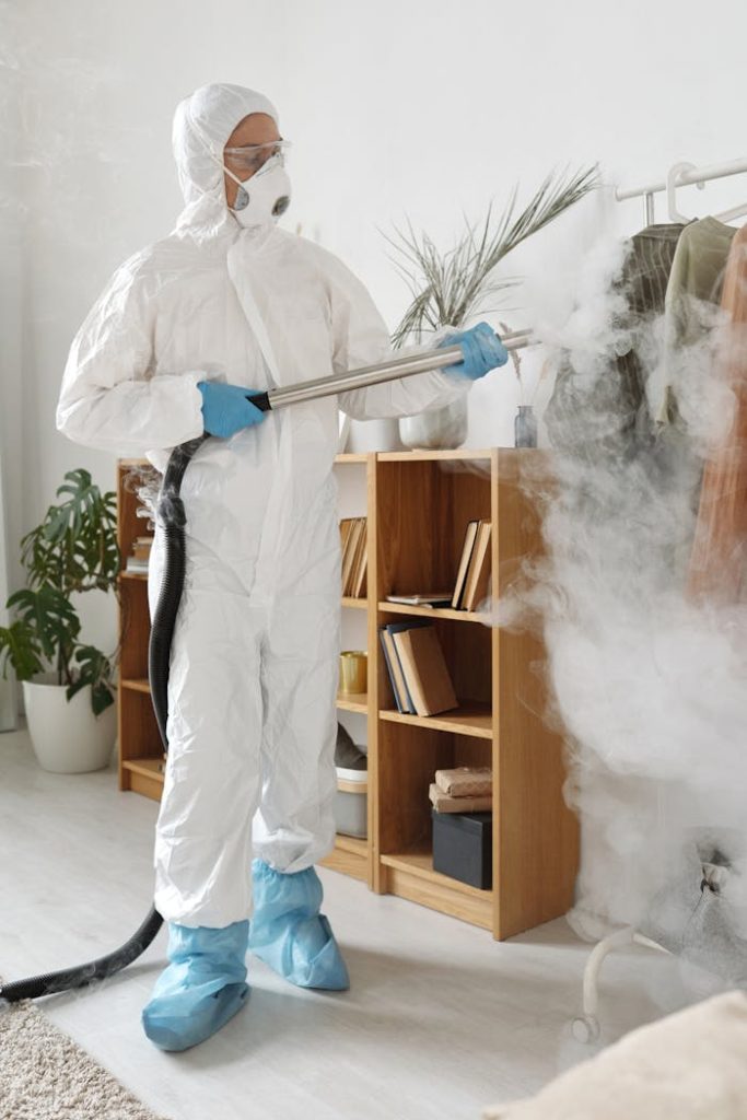An adult wearing PPE sprays disinfectant indoors, combating Covid-19.