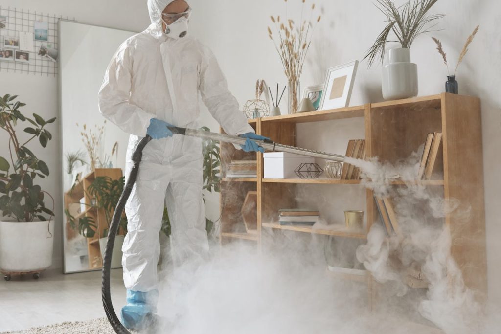 A professional in PPE, disinfecting a wooden shelf indoors during daylight.